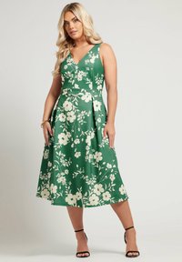 Vihreä kukkamekko, jossa on V-pääntie, olkaimeton malli, jossa on vohvelivärisiä kukkia, liehuva hame ja sileä kangastekstuuri. Mustat remmijalat.