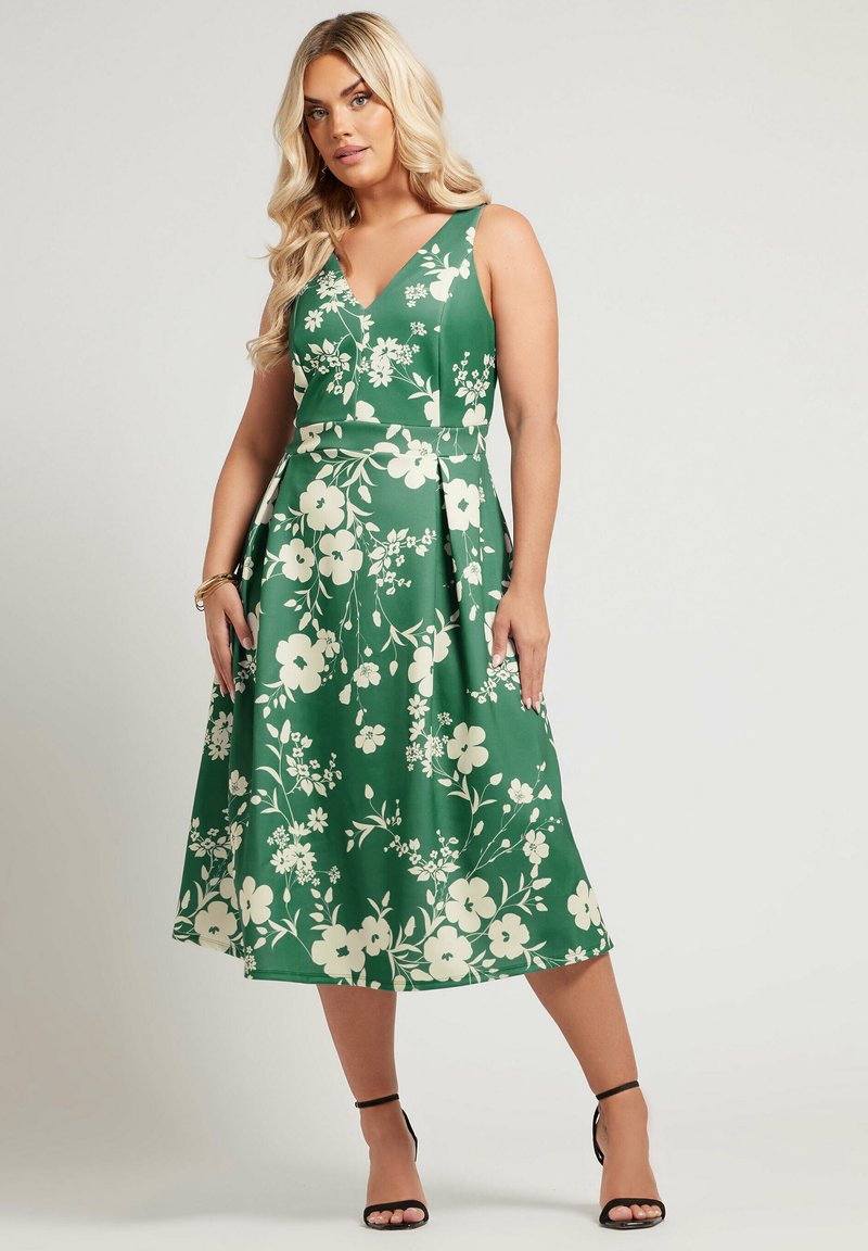 Vihreä kukkamekko, jossa on V-pääntie, olkaimeton malli, jossa on vohvelivärisiä kukkia, liehuva hame ja sileä kangastekstuuri. Mustat remmijalat.