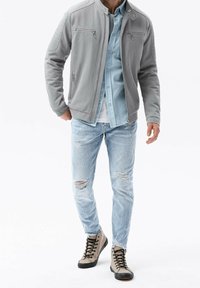 Grijze textuurjack met een opstaande kraag, gelaagd over een lichtblauw overhemd met knopen, gecombineerd met versleten lichtblauwe jeans en beige hoge laarzen.