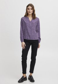 Lila långärmad blus med vita prickar, knäppning framtill och rynkade manschetter. Kombinationen är kompletterad med svarta slim fit-jeans och svarta loafers.
