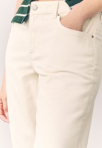 Pantalon en coton couleur crème avec une coupe droite, doté d'une poche avant et d'une fermeture à bouton visible. Texture subtile avec une quincaillerie minimale.