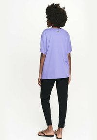 Liliowa koszula z krótkim rękawem o luźnym kroju, zestawiona z dopasowanymi czarnymi spodniami. Prosty design, miękka faktura i minimalne detale na plecach.