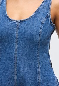 Κοντινό πλάνο ενός ατόμου που φοράει εφαρμοστό μπλε τζιν φόρεμα χωρίς μανίκια με λαιμόκοψη scoop και λεπτομέρεια πλευρικού φερμουάρ.