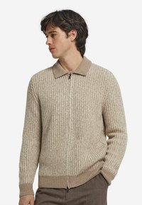 Beige ribbad stickad cardigan med dragkedja och en kontrasterande krage. Har texturerad tyg och en figursydd midjedetalj.