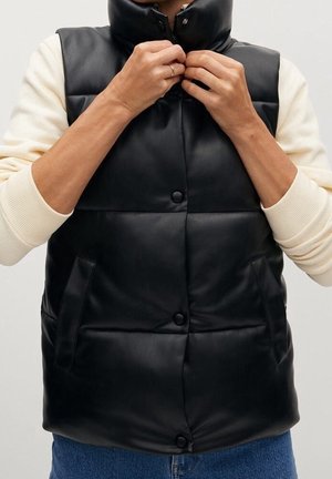 Henkilö kiinnittää nappeja mustassa topatussa hihattomassa liivissä, joka on päällä kermaisenvärisen pitkähihaisen paidan ja sinisten farkkujen päällä.