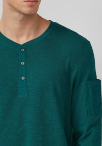 Man in een teal lange mouwen henley shirt met drie zwarte knopen en een klein zakje op de linkerbovenarm.