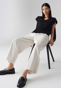 Top nero a maniche corte con scollo a V, pantaloni a righe crema a gamba larga e mocassini neri. Modella seduta su una sedia nera contro uno sfondo chiaro.