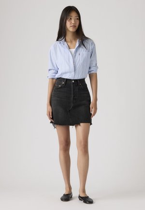 Kvinde iført en lyseblå stribet skjorte med knapper, sort flosset denimnederdel og sorte slip-on sko, stående mod en ensfarvet baggrund.