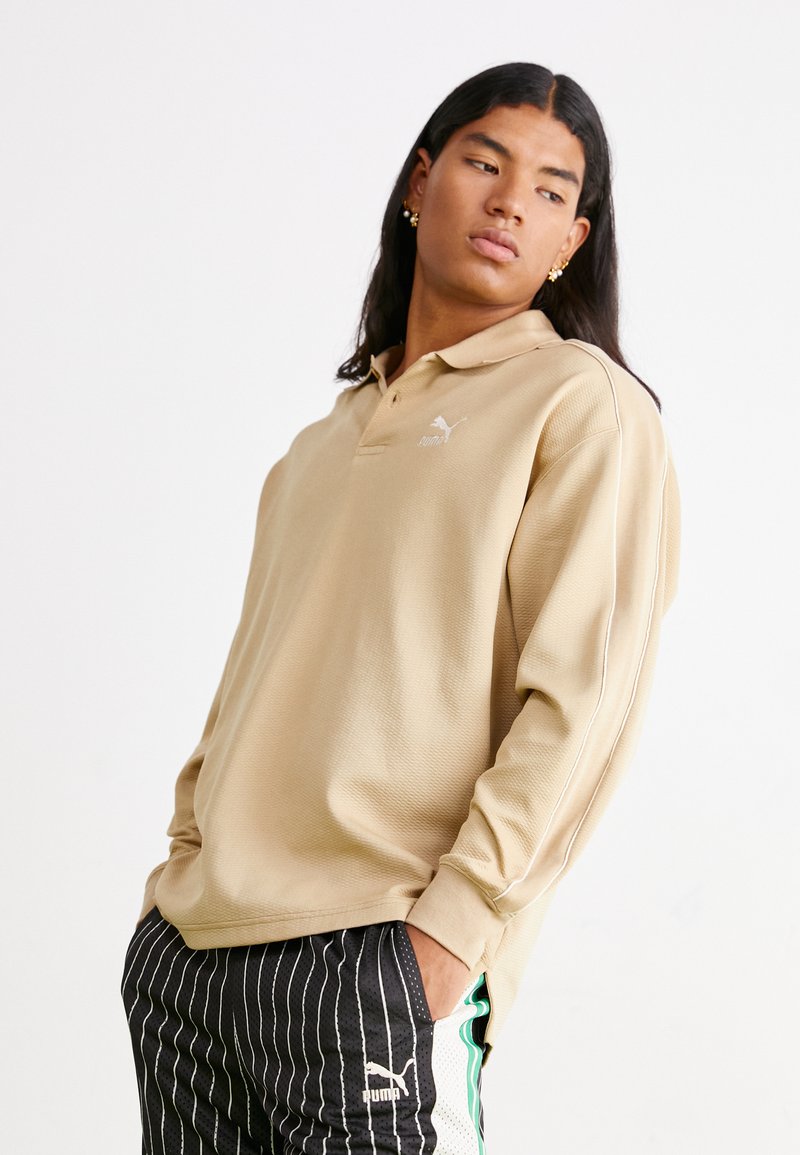 Beige Polohemd mit langen Ärmeln aus strukturiertem Stoff, mit einem weißen Logo und seitlichen Streifen. Getragen mit schwarz-weißen gestreiften Hosen.