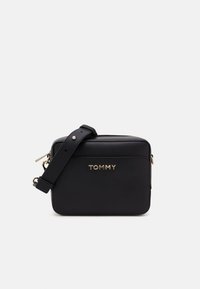 Czarna syntetyczna torebka na ramię w prostokątnym kształcie, z złotym logo "TOMMY", zamknięciem na zamek błyskawiczny i regulowanym paskiem. Gładka faktura na całej powierzchni.