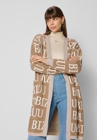 Lange cardigan in tan breiwerk met witte letters, open voorkant en geribbelde manchetten. Gepaarde met lichtgewassen jeans en een beige top.