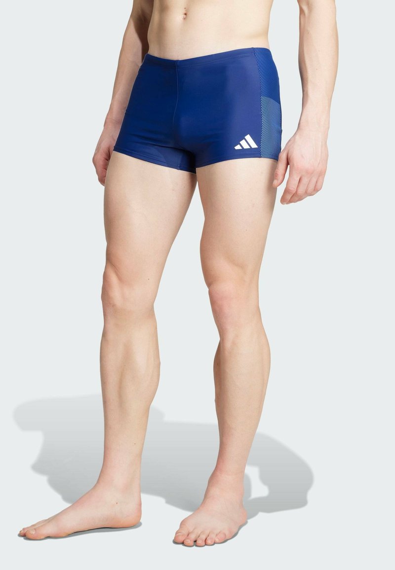 Bañador corto azul marino con textura suave, que presenta un panel de malla contrastante en un lado y un logo de adidas en la parte delantera.