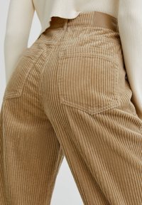 Pantalon en velours côtelé beige avec une texture côtelée verticale, doté de poches arrière et d'une étiquette en cuir sur la ceinture.