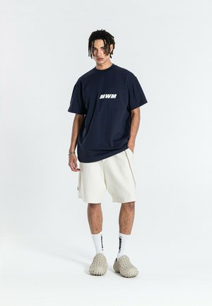 Joven con rastas lleva una camiseta azul marino oversize con el logo "MWM", pantalones cortos blancos, calcetines altos blancos y zapatos beige con textura sin cordones.