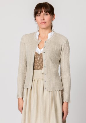 Frau mit braunen Haaren, die eine beige strukturierte, geknöpfte Strickjacke über einer weißen Bluse und einem beige gemusterten Kleid vor einfarbigem Hintergrund trägt.