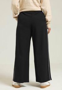 Svarta vida byxor med en slät finish, som har vita sidostråk och resårmidja, kombinerat med ljusa sneakers.