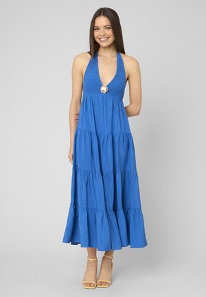 Jeune femme souriante, portant une robe longue à volants bleue avec un détail coquillage au niveau de la poitrine et des talons jaunes à bride cheville, debout sur fond gris.