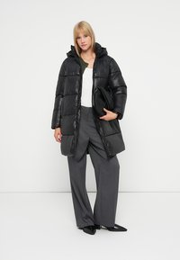 Casaco acolchoado preto com capuz, design matelassé e textura brilhante. Usado sobre uma blusa branca e calças cinzentas, acompanhado de uma bolsa preta.