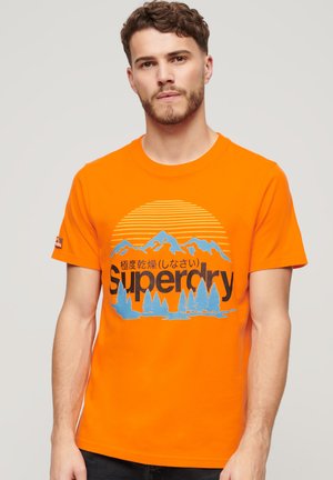 Camiseta de algodón naranja con un diseño gráfico de montañas y árboles en azul y negro. Cuello redondo acanalado y mangas cortas.