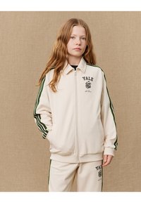Kremfarget zip-up jakke med grønne striper på ermene, med "YALE" logo og grafisk trykk. Mykt stoff, avslappet passform, sammen med matchende bukser.