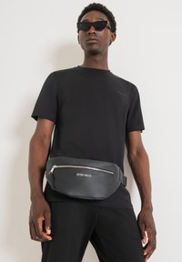 Marsupio in pelle nera con superficie testurizzata e zip in argento. Indossato in vita, presenta il logo "ANTONY MORATO" in bianco.