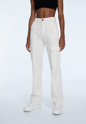 Woman wearing high-waist white cargo pants, black cropped top, and white sneakers standing against plain background.