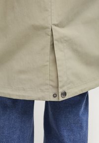 Khaki buitenkleding met een rechte snit, zijschade en metalen drukknoopdetails, gecombineerd met donkerblauwe denim jeans. Zachte, gladde stoftextuur.