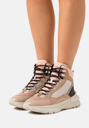 Beige-rosa Calvin Klein Jeans High-Top-Sneaker mit schwarzen Schnürsenkeln und klobigen weißen Sohlen, getragen an nackten Beinen.