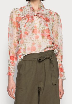 Mannequin portant un chemisier crème transparent à imprimé floral corail et un nœud au cou, associé à un pantalon olive noué à la taille.