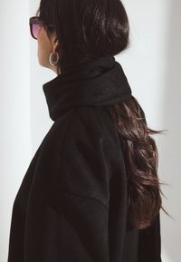 Czarny wełniany płaszcz z wysokim kołnierzem, miękką teksturą i luźnym krojem. Długie, falowane włosy częściowo widoczne; nosi oversize'owe okulary przeciwsłoneczne i srebrne kolczyki w kształcie kół.