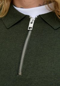 Mørkegrøn strikket pullover med lynlås foran. Har en krave og sølv lynlåsdetaljer, der viser et struktureret stof.