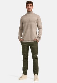 Beige kötött garbó pulóver texturált csíkokkal, olíva zöld slim-fit nadrággal és világos bézs sportcipővel párosítva.