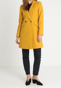 Dubbelzijdige gele jas gemaakt van wolmix, met zijzakken en scherpe revers, gecombineerd met zwarte jeans en puntige zwarte hakken.