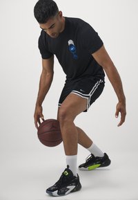 Schwarzes Nike-T-Shirt mit Logo, schwarze Shorts mit weißen Streifen, Basketball in der Hand, schwarz-grüne Sportschuhe, weiße Socken, konzentrierte Haltung.