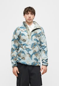 Νέος άνδρας που φοράει μπλε και μπεζ φλις πουλόβερ με μοτίβο και μισό φερμουάρ, μαζί με μαύρο παντελόνι cargo, στέκεται μπροστά σε ένα απλό λευκό φόντο.