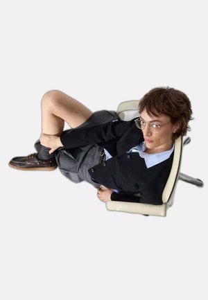 Persona con i capelli corti che indossa occhiali, cardigan nero, camicia blu, gonna grigia e scarpe marroni, seduta di lato su una sedia da ufficio bianca.