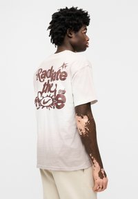 Μπεζ βαμβακερό t-shirt με μεγάλη, στιλιζαρισμένη γραφική απεικόνιση "Radiate the" σε καφέ χρώμα στην πλάτη. Κοντά μανίκια και άνετη εφαρμογή.