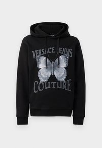 Hanorac cu glugă de culoare neagră, realizat din bumbac, având un grafic mare cu un fluture gri și textul "VERSACE JEANS COUTURE" scris cu litere îngroșate.