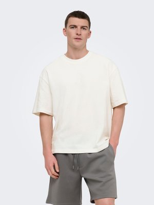Jeune homme portant un t-shirt blanc ample à manches courtes et un short gris à cordon, debout avec une main dans la poche.