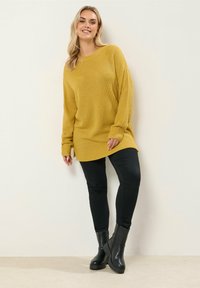 Pull en maille jaune moutarde à coupe décontractée, manches longues, fentes latérales ; associé à un pantalon noir ajusté et des bottines noires.