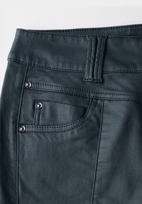 Tela de denim verde oscuro con una textura suave, presenta una cinturilla, un bolsillo trasero con remaches de metal y detalles de costura en contraste.