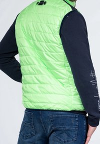 Pikowana kamizelka w jaskrawej neonowej zieleni z pikowaną fakturą, zestawiona z granatową koszulą z długim rękawem. Zapięcia na kołnierzu z czarnymi guzikami akcentowymi.