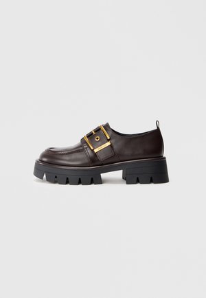 Scarpa in pelle marrone con suola nera spessa, presenta una grande fibbia dorata e una texture liscia, progettata con punta arrotondata.