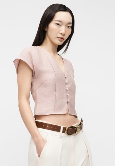 Γυναίκα που φοράει ανοιχτό ροζ κοντομάνικο κοντό μπλουζάκι με κουμπιά, λευκό ψηλόμεσο παντελόνι και καφέ ζώνη με χρυσή αγκράφα.