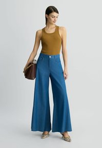 Geripptes senfgelbes Tanktop, hoch taillierte, blaue weit geschnittene Jeanshose und bedruckte spitze Pumps, kombiniert mit einer braunen Schultertasche.