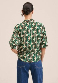 Chemise à manches courtes beige avec un motif géométrique abstrait vert et marron, dotée d'un col et de manches légèrement bouffantes.