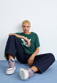 Person with short blonde hair sitting on floor wearing green Adidas shirt, wide-leg dark jeans, and pale purple sneakers with white soles.