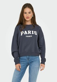 Giovane donna con lunghi capelli castani che indossa un felpa blu navy con la scritta "PARIGI FRANCIA" e jeans azzurri, in piedi contro uno sfondo uniforme.