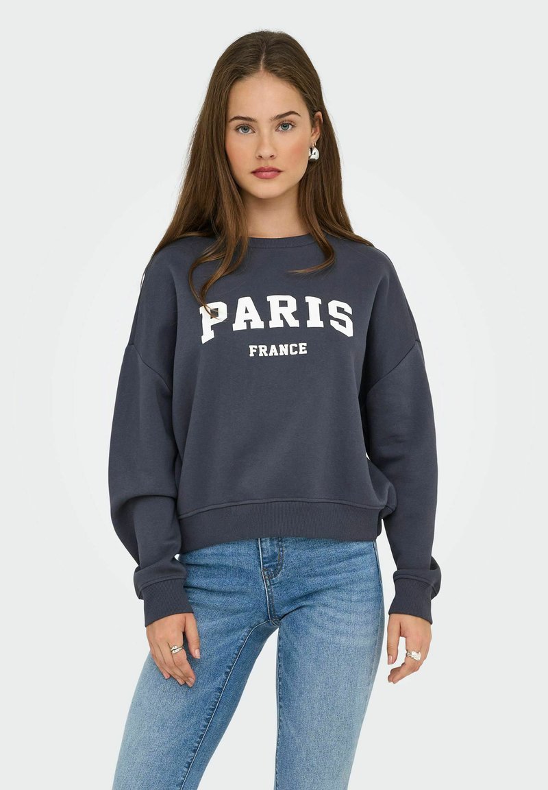 Giovane donna con lunghi capelli castani che indossa un felpa blu navy con la scritta "PARIGI FRANCIA" e jeans azzurri, in piedi contro uno sfondo uniforme.