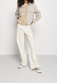 Beige zip-up jacka, zebra-mönstrad topp i neutrala toner och vida vita byxor, tillsammans med vita sneakers med gröna detaljer.